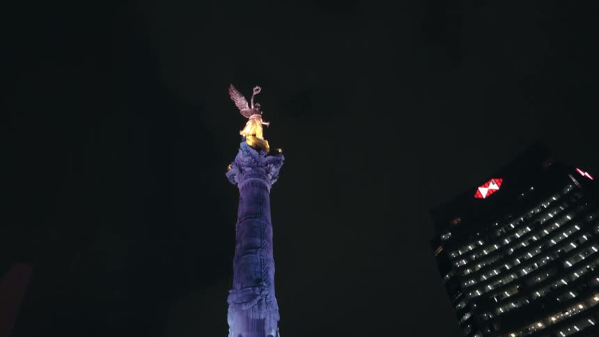 A low-angle tilt-up reveals the Angel of Independence monument illuminated at night against the backdrop of a modern skyscraper in Mexico City