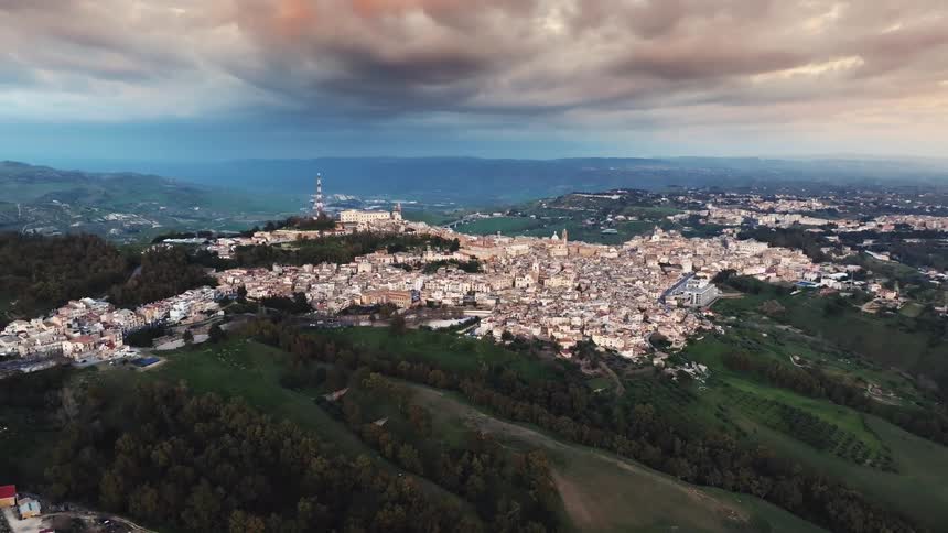 A cinematic aerial shot slowly pulls back to reveal a sprawling historic town on a verdant hillside under a dramatic, colorful sky