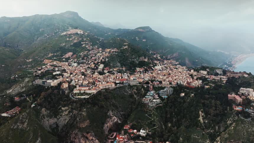 A stunning aerial view of the historic town of Taormina, built into the lush green mountainside overlooking the Italian coast