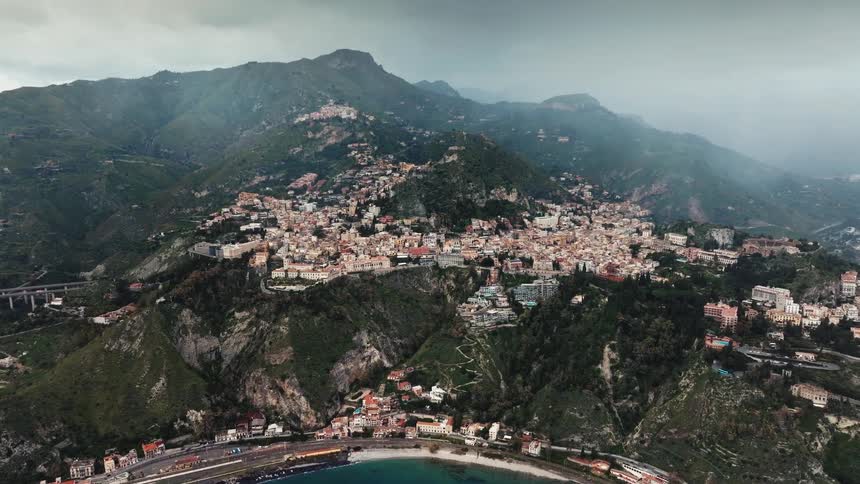 A dramatic aerial view pushes slowly towards the beautiful coastal town of Taormina, nestled in the mountains of Sicily on a cloudy day