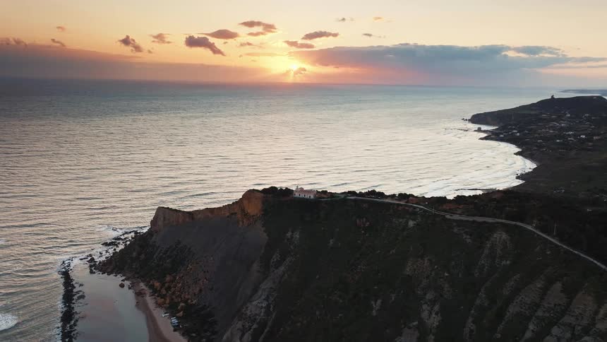 A serene aerial view shows the sun setting over the ocean, illuminating a rugged green cliff and a solitary house on the coastline