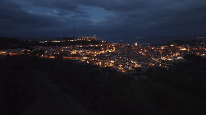 A stunning aerial view slowly pulls back to reveal the illuminated historic city of Caltagirone, Sicily, under a dark, cloudy night sky