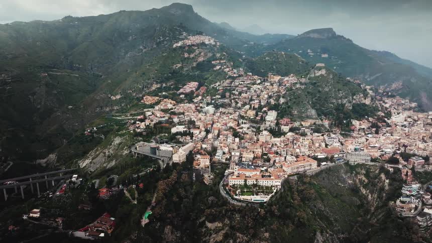 An epic aerial view slowly reveals the historic town of Taormina, Sicily, built into the side of a lush green mountain on an overcast day