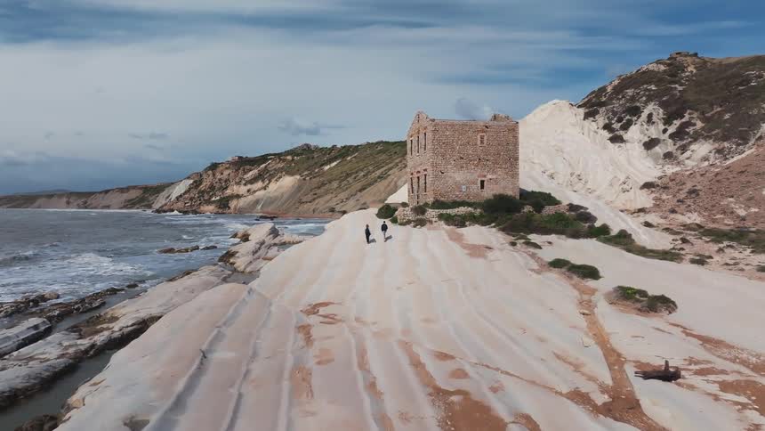 Aerial view of two people walking along a unique white rock coastline towards an ancient ruined building by the sea