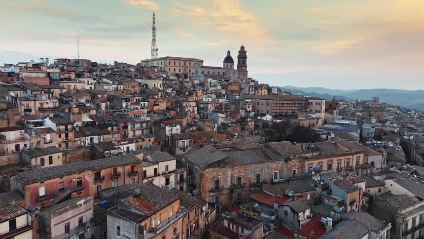A slow aerial dolly shot over the historic rooftops of Caltagirone, a hilltop town in Sicily, Italy, during a beautiful sunset