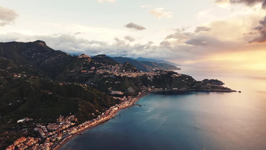 An epic aerial shot showcases the stunning Italian coastline, with a town clinging to the mountainside as the sun sets over the calm sea