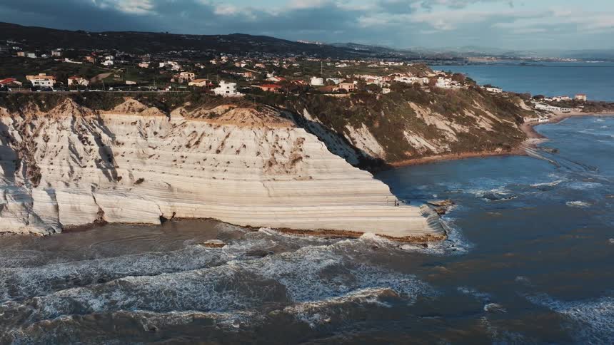 A stunning aerial view shows the Scala dei Turchi's white cliffs in Sicily as waves crash against the shore below a coastal town
