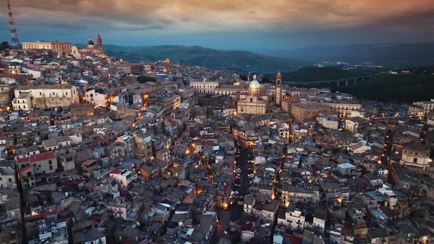 An aerial view of the historic city of Caltagirone in Sicily, Italy, as its lights begin to glow at twilight against a dramatic sky