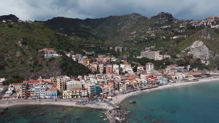 A stunning aerial view pulls back from a colorful Italian coastal town nestled between lush green mountains and the turquoise Mediterranean Sea under dramatic clouds