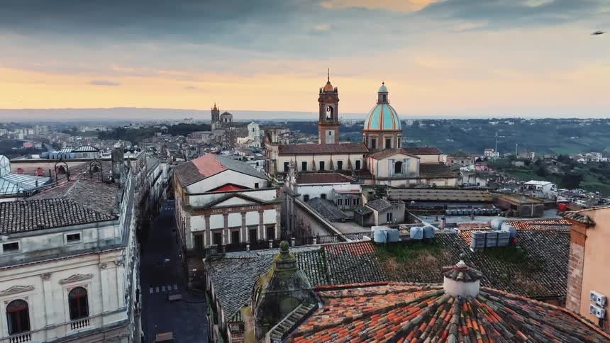 A slow aerial push over the historic rooftops of an ancient Sicilian city towards a majestic cathedral at sunset