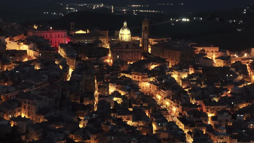 An enchanting aerial view zooms in on a historic Italian city at night, its ancient buildings and cathedral glowing with warm city lights