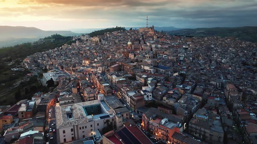 Stunning aerial pull-back shot reveals the historic hilltop city of Caltagirone in Sicily, Italy, as the lights turn on at sunset