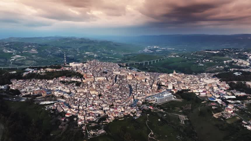 A majestic aerial shot slowly reveals a historic city nestled among rolling green hills in Sicily, Italy, under a dramatic, cloudy sky at dusk