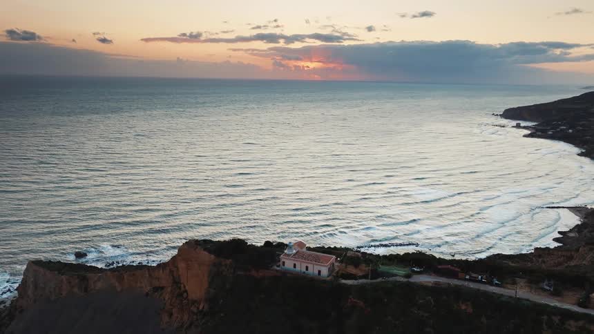 An aerial drone shot flies over a picturesque coastline at sunset, revealing a lone building on a dramatic cliff overlooking the gentle ocean waves