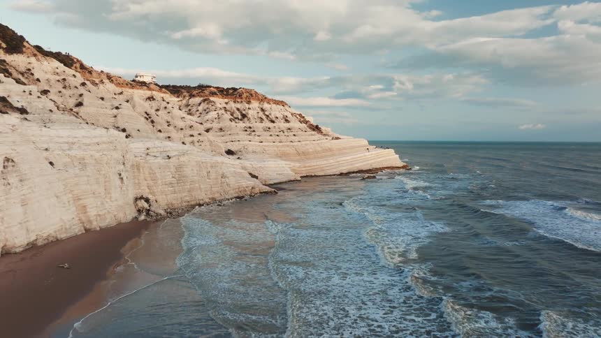 An aerial view reveals the stunning Scala dei Turchi, where white marl cliffs meet the gentle waves of the sea under a cloudy sky