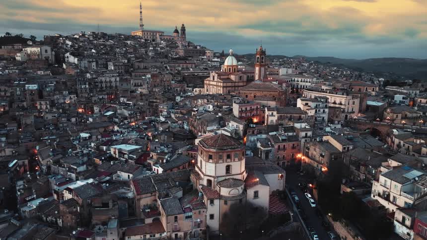 A stunning aerial view captures the ancient Sicilian city built on a hill, its beautiful architecture illuminated at twilight under a golden sunset sky
