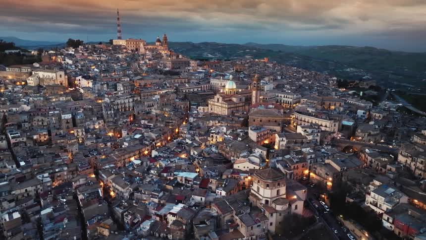 An expansive aerial shot captures the historic city of Caltagirone in Sicily as evening lights begin to glow under a beautiful sunset