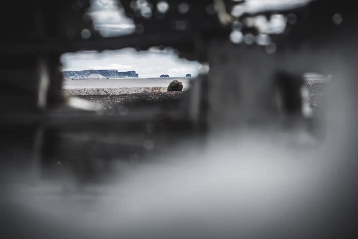 A slow focus pull reveals a desolate black sand beach and ocean seen through the wreckage of a crashed plane