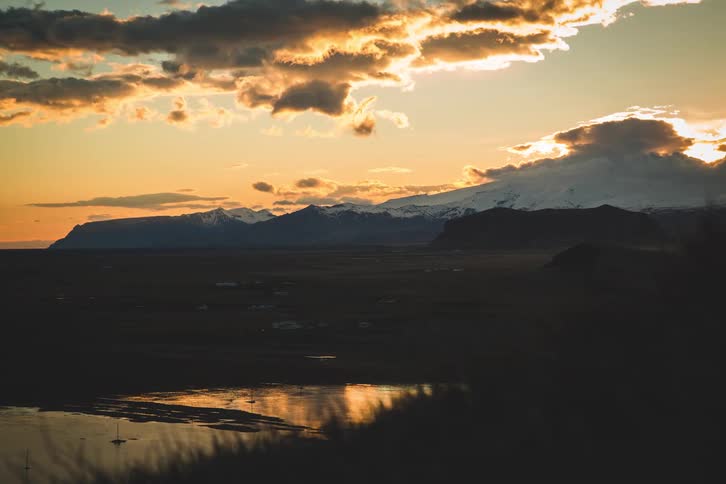 A stunning timelapse of the sun setting behind snow-capped mountains, casting a golden light over a vast valley and a calm body of water