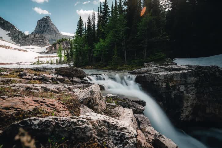 A majestic timelapse captures clouds rolling over snow-covered mountains while a crystal clear river cascades down a rocky waterfall