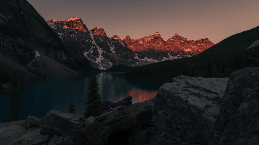 A beautiful timelapse captures the sunrise alpenglow on the snow-covered mountains surrounding a tranquil lake