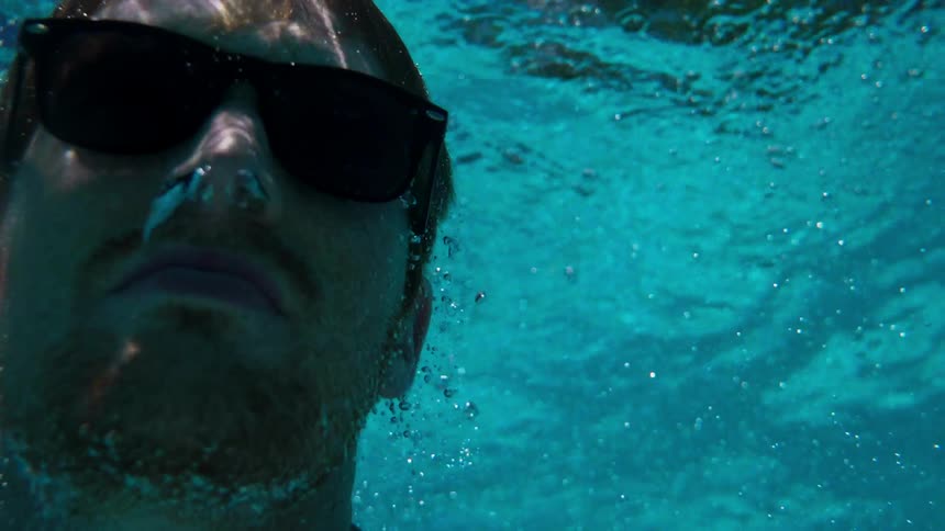A man wearing dark sunglasses is floating and goes to the surface of the water
