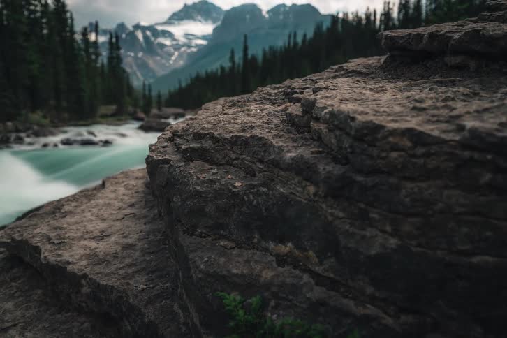 A slow-motion timelapse reveals a stunning mountain landscape with a turquoise river flowing through a pine forest towards snow-capped peaks