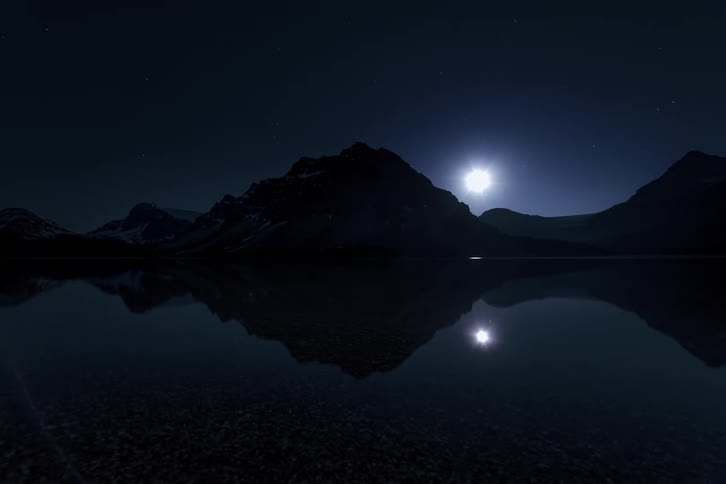 A time-lapse shows the moon rising from behind a silhouetted mountain range, perfectly reflected in a still lake under a starry night sky