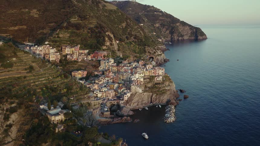 An aerial drone shot pulls back to reveal the beautiful Italian coastal village of Manarola in Cinque Terre nestled on a cliffside at sunrise