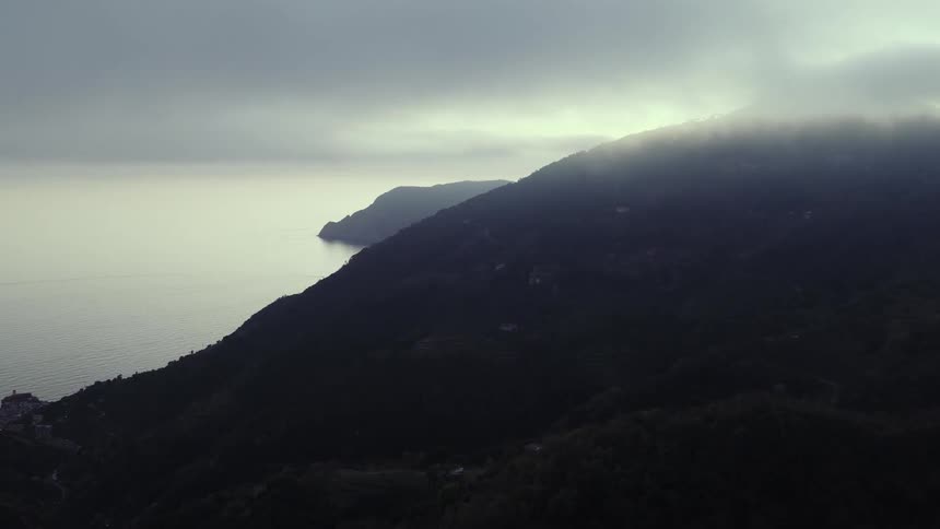 A slow aerial shot pushes in over a moody coastal mountain range shrouded in fog and low clouds at dawn