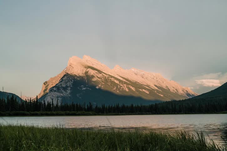 A stunning sunset timelapse shows golden light turning to pink alpenglow on a majestic mountain peak over a calm lake