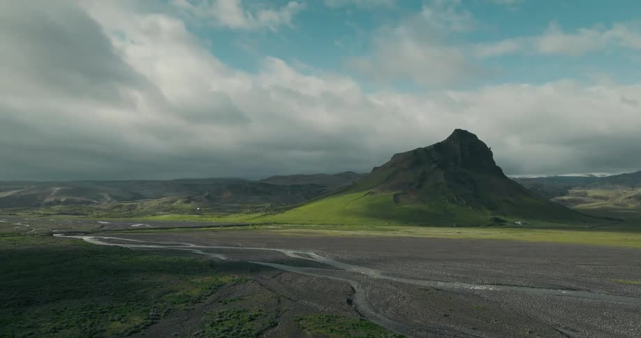 A wide, static shot captures a majestic green mountain rising from a vast river valley under a dramatic cloudy sky in Iceland