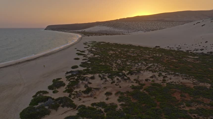 A serene aerial view captures a vast sandy coastline where the ocean meets rolling dunes under the golden light of sunset