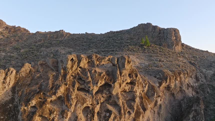 Aerial drone footage pulls back from a magnificent natural rock arch formation on a rugged mountainside during a serene golden hour
