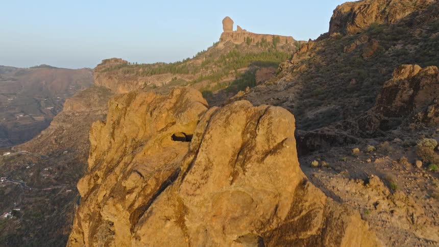A stunning aerial view ascends a golden cliff face to reveal the iconic Roque Nublo landmark in Gran Canaria at sunset