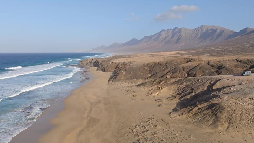 An epic aerial view reveals a vast, remote coastline with ocean waves crashing against a sandy beach, overlooked by a camper van and distant mountains