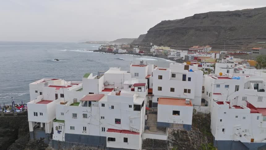 An aerial shot slowly rises to reveal a beautiful white coastal village built precariously on a volcanic cliff by the ocean