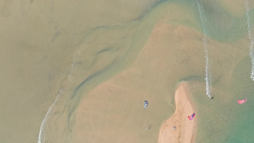 An aerial top-down view of kitesurfers gliding across a serene sandbar in shallow, clear water