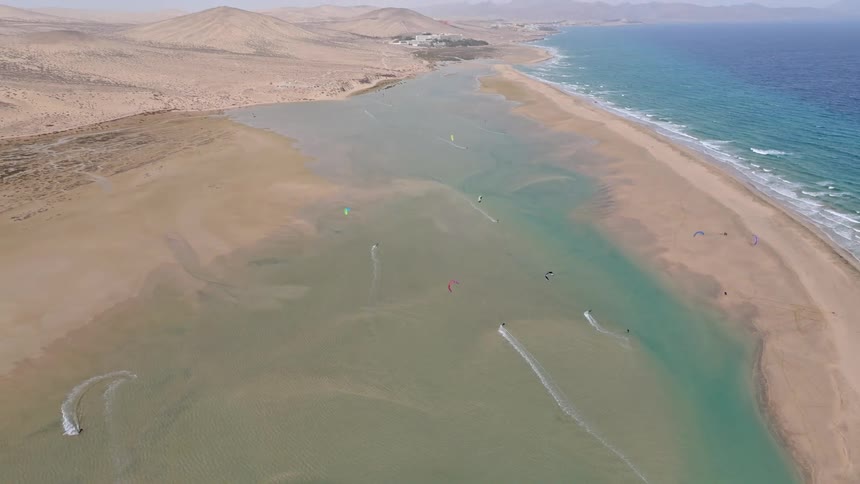A wide aerial shot reveals numerous kitesurfers enjoying the shallow turquoise lagoon on a vast desert coastline