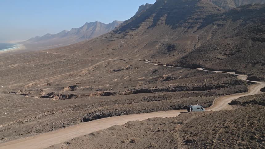 Aerial view of a camper van navigating a winding dirt road through a vast, mountainous desert landscape towards the coast