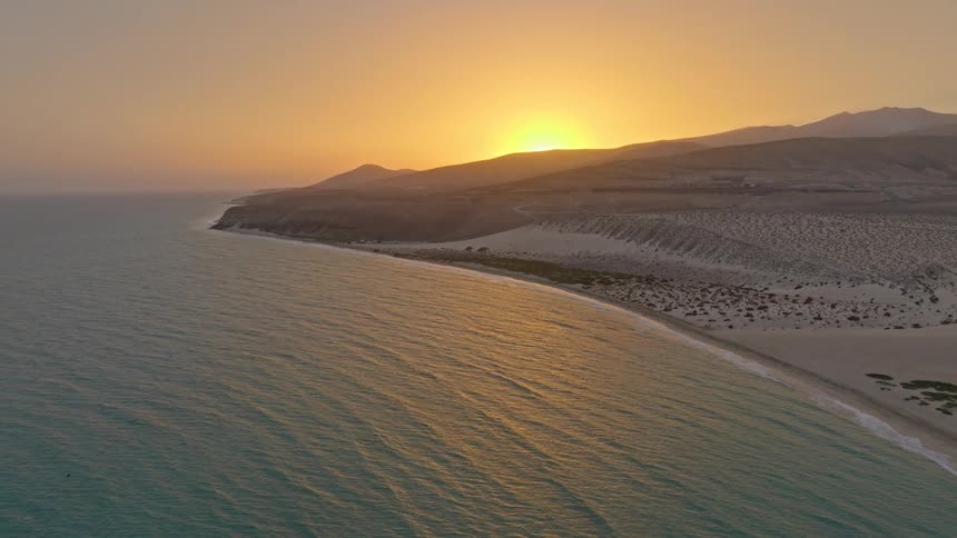 A serene aerial shot captures the golden sunset reflecting on the calm ocean along a vast desert beach