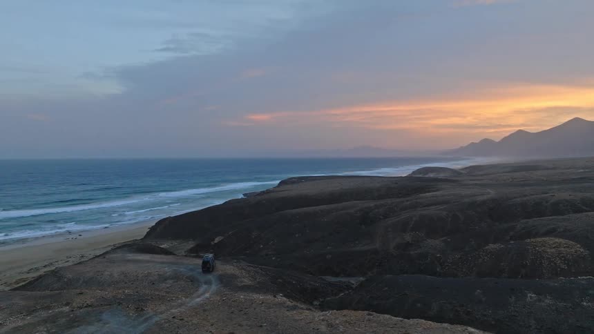Aerial view of a lone camper van parked on a remote, volcanic coastline overlooking the ocean at sunset