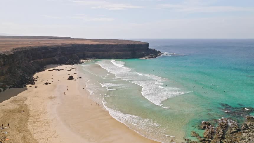Aerial drone shot flies over a cliffside with camper vans to reveal a stunning turquoise beach cove with surfers