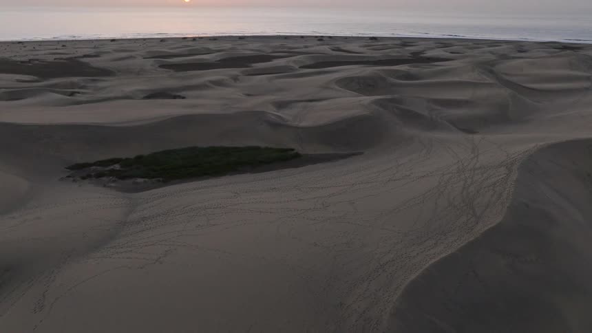 An aerial view of vast sand dunes slowly tilts up to reveal a stunning sunrise over the distant ocean horizon