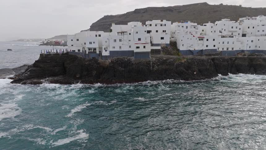 Dramatic aerial view descending towards a unique white village built on a rocky peninsula as ocean waves crash against the cliffs