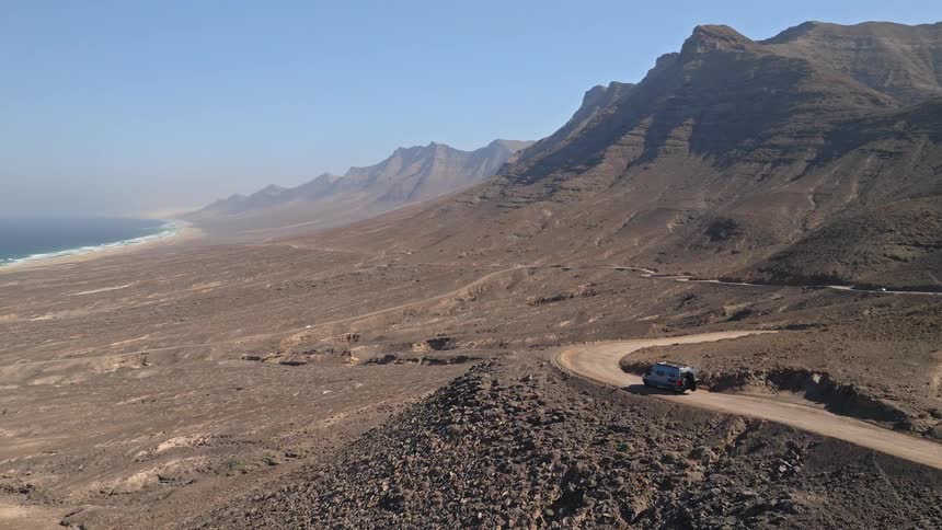 Scenic aerial view of a camper van driving along a winding cliffside dirt road in a vast, arid mountain landscape overlooking the ocean