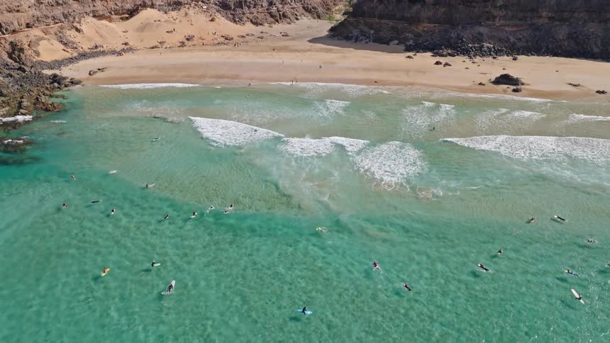 An aerial view tilts up to reveal surfers in vibrant turquoise waters at a secluded beach cove framed by dramatic arid cliffs
