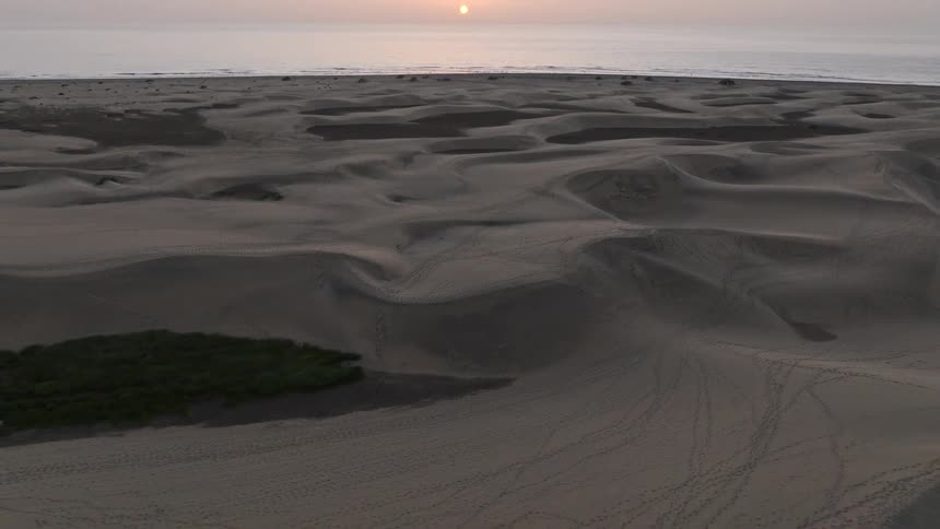 A stunning aerial view pulls back over vast sand dunes to reveal a serene sunrise over the ocean horizon