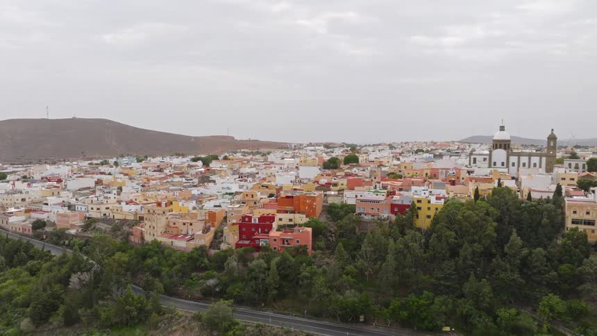 An ascending aerial view reveals the historic and colorful town of Agüimes in Gran Canaria, nestled in the hills under an overcast sky