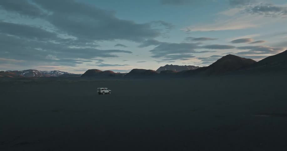 An epic, wide, static shot shows a lone white off-road vehicle parked in the vast, empty black sand desert of the Icelandic highlands